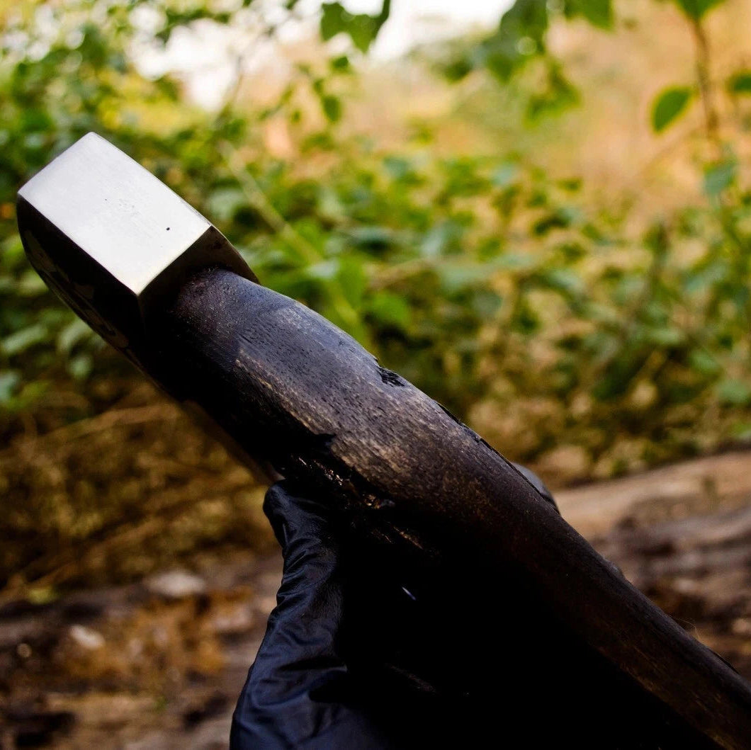 Wooden axe handle with a metallic head held by a gloved hand against a blurred natural background