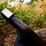 Wooden axe handle with a metallic head held by a gloved hand against a blurred natural background